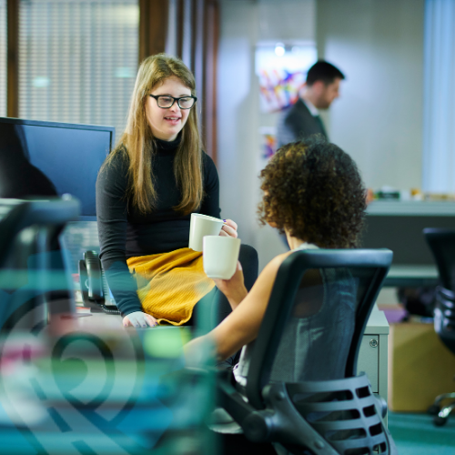 Two women having a relaxed, friendly conversation in a bright office space — representing positive workplace culture, connection, and employee wellbeing.